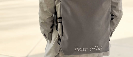 Close-up of a gray backpack embroidered with the words 'hear Him' on a man's back
