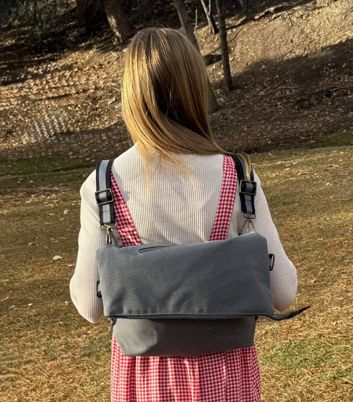 Girl with a checkered dress carrying a folded gray backpack in an outdoor setting