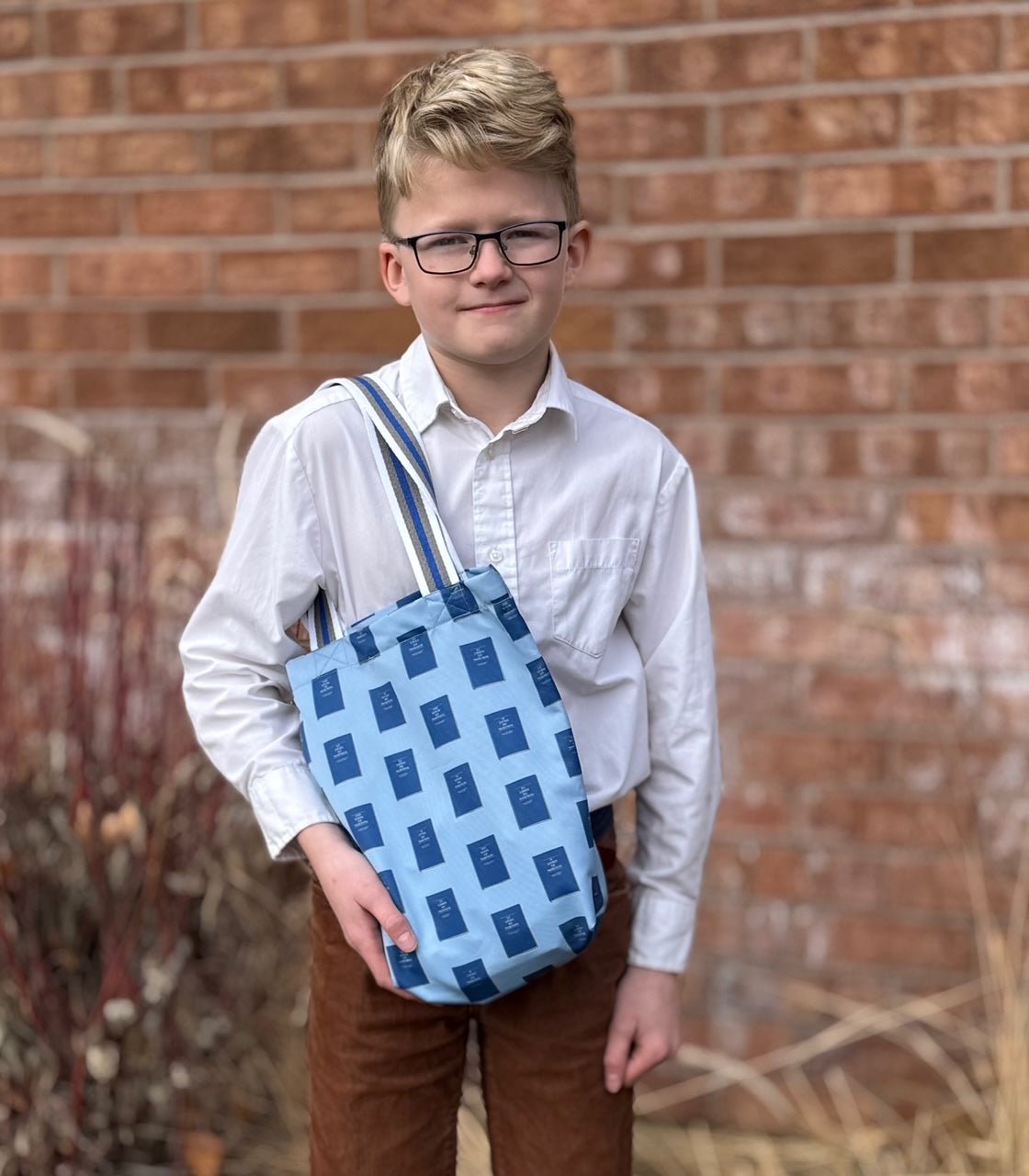 A blue tote bag with a repeating pattern of Books of Mormon. The bag has a striped shoulder strap with white, blue, and gray stripes. The bag is held by a young boy wearing a white button-down shirt, glasses, and brown pants.
