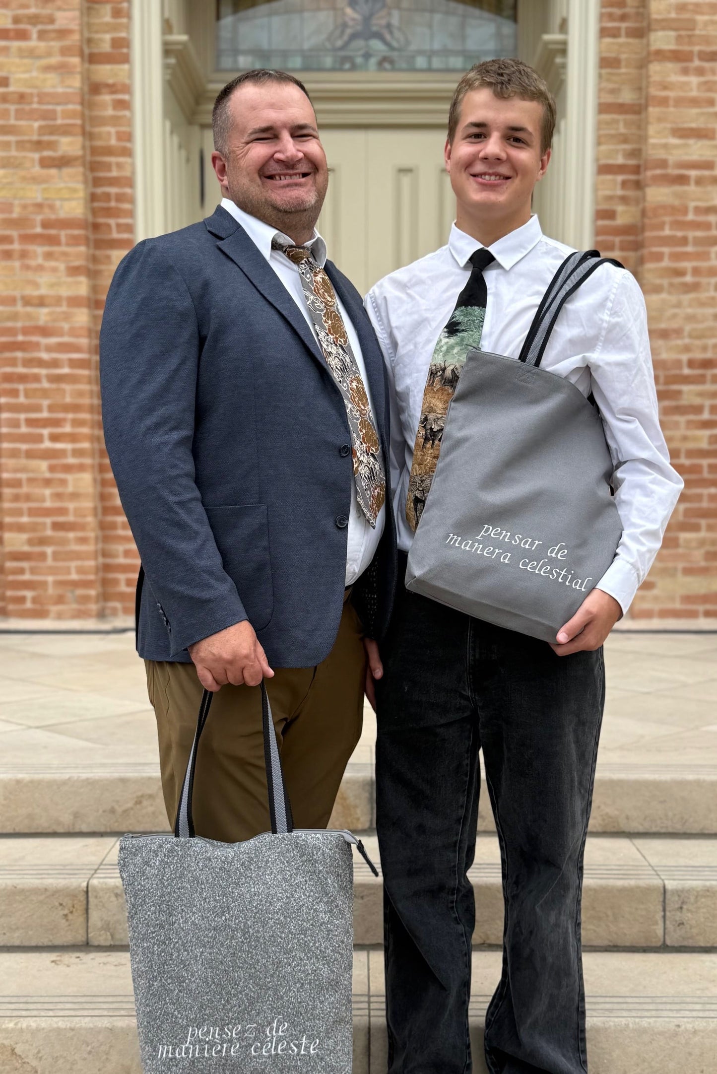 Father and son smiling on temple steps, holding temple bags with “pensez de manière céleste” and “pensar de manera celestial” embroidered on front