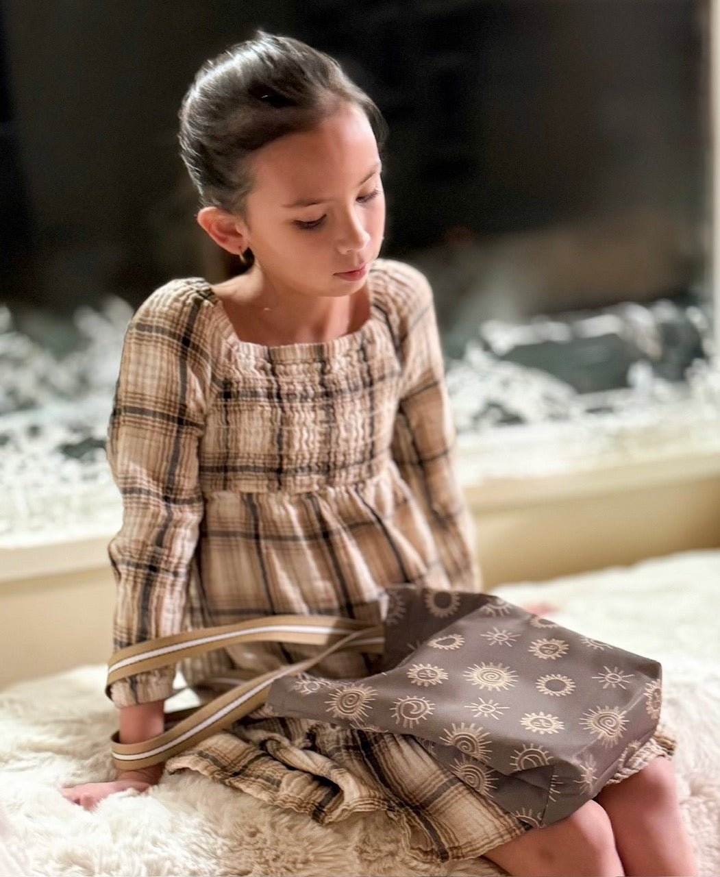 A young girl wearing a plaid dress with a square neckline. The dress is in shades of brown and tan. A brown tote bag with a sunstones pattern is held in her lap.