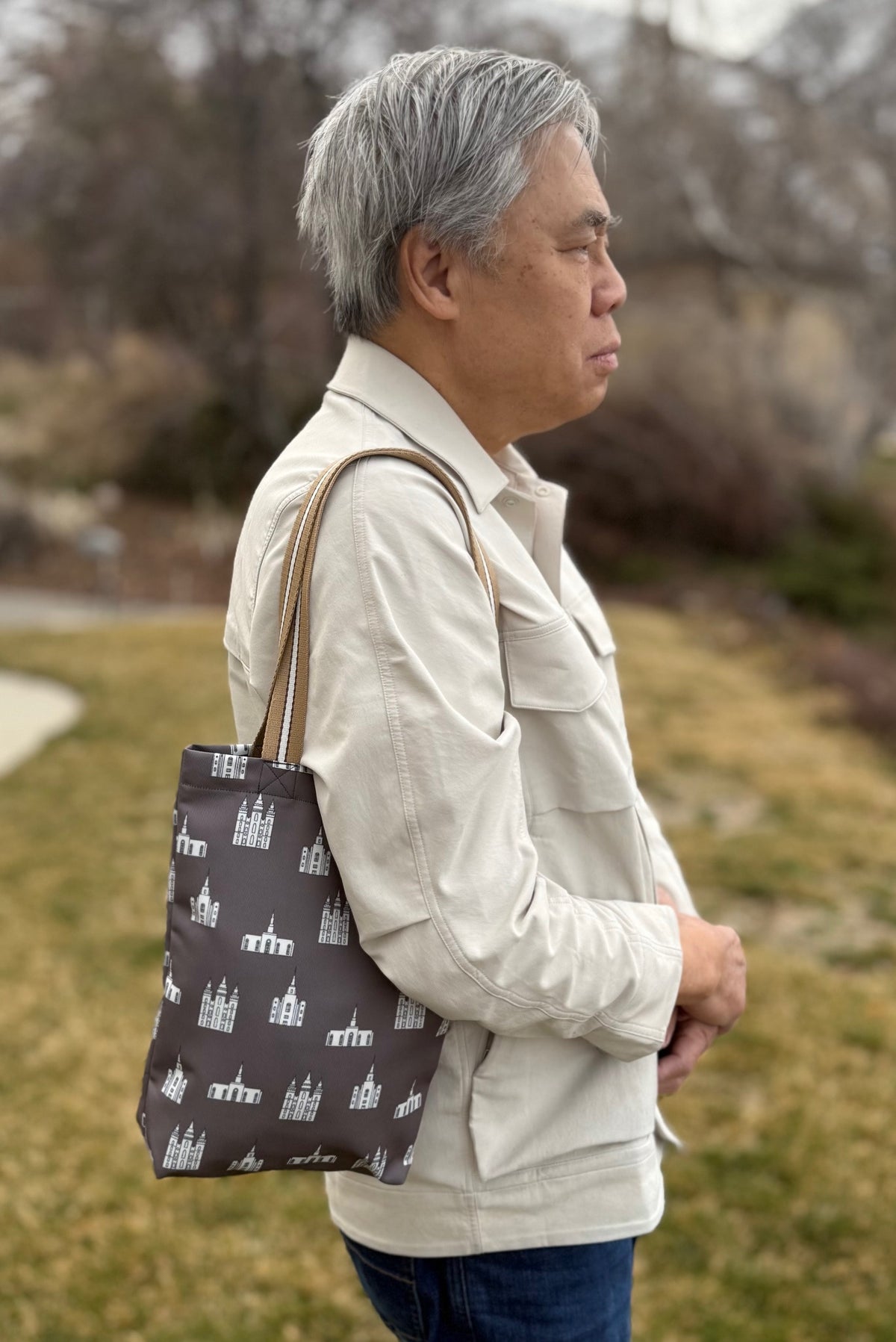 A brown tote bag featuring a repeating pattern of white temple illustrations. The bag has a beige and white striped strap and is carried over the shoulder by an older man with a light jacket.