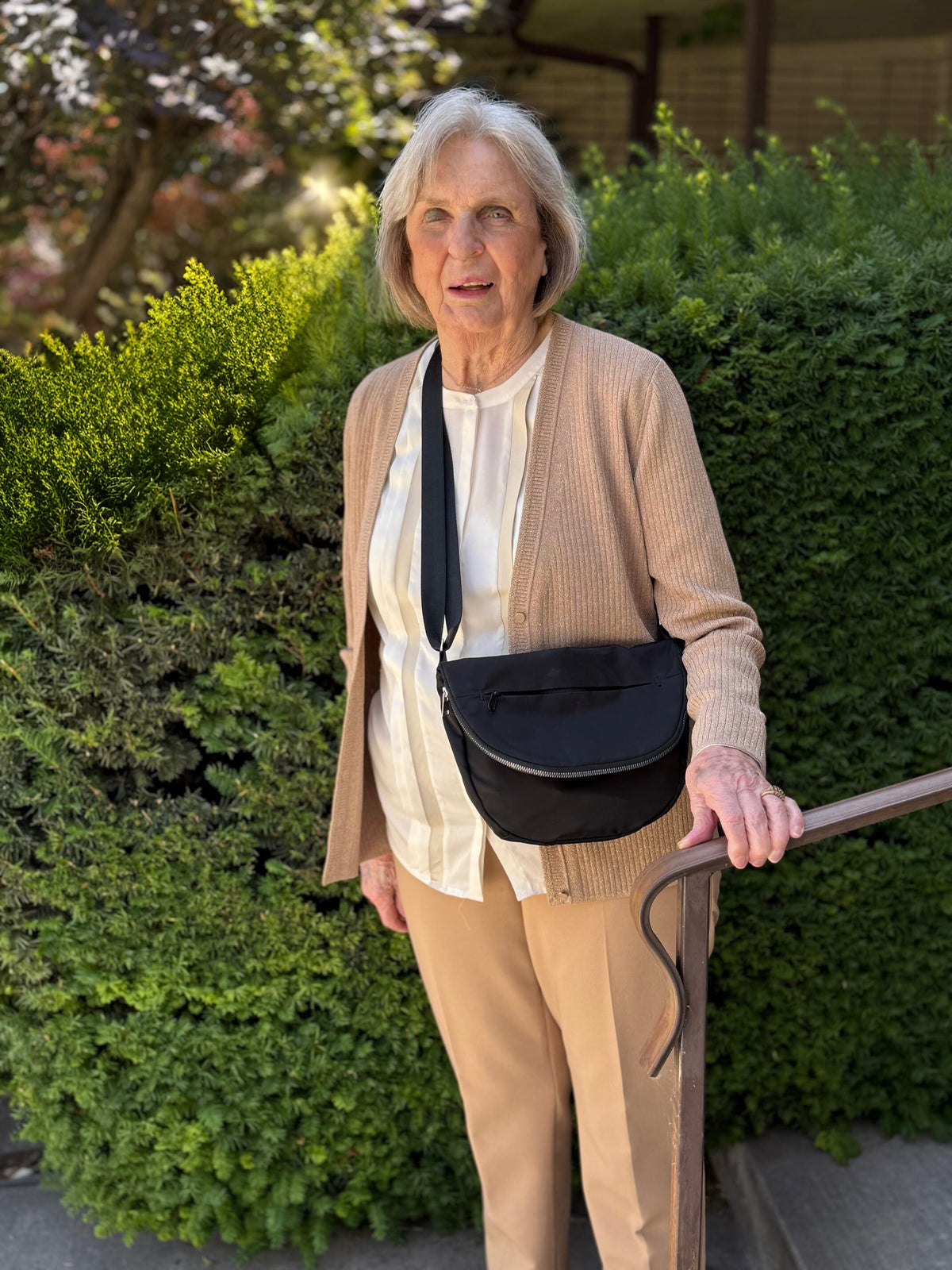 A woman wearing a tan cardigan, a cream-colored blouse, and tan pants is wearing a black crossbody bag. The background includes green bushes and a metal railing.