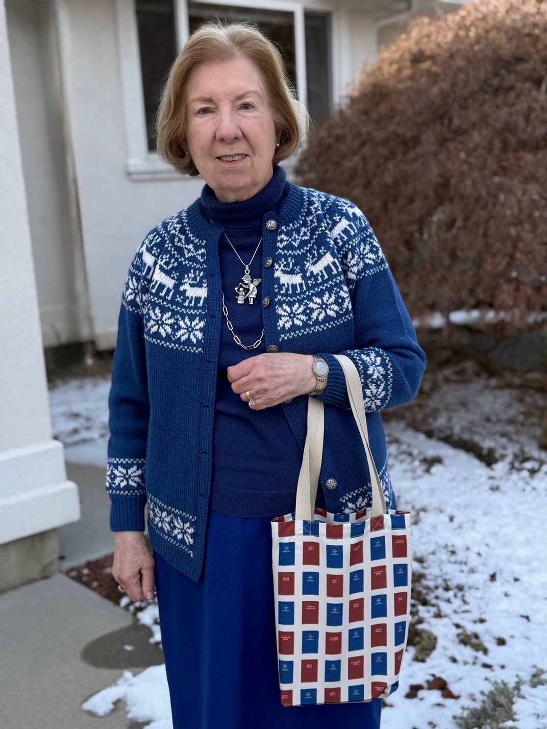 A woman wearing a navy blue sweater and cardigan with a white snowflake and reindeer pattern. She is holding a tote bag with a pattern of red Bibles and blue Books of Mormon.