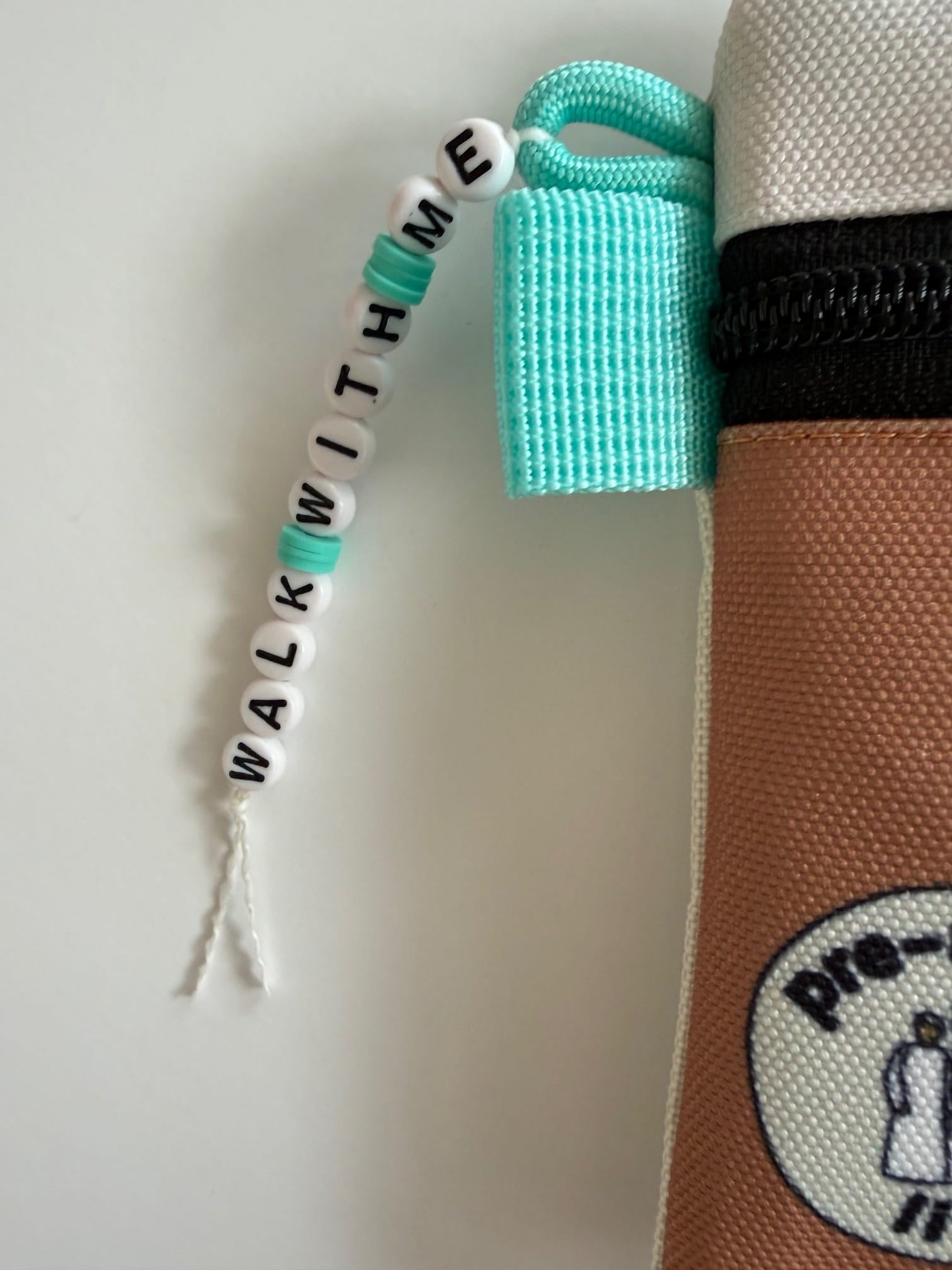 Close up of charm with letter beads spelling 'WALK WITH ME' attached to All-In Pouch strap 