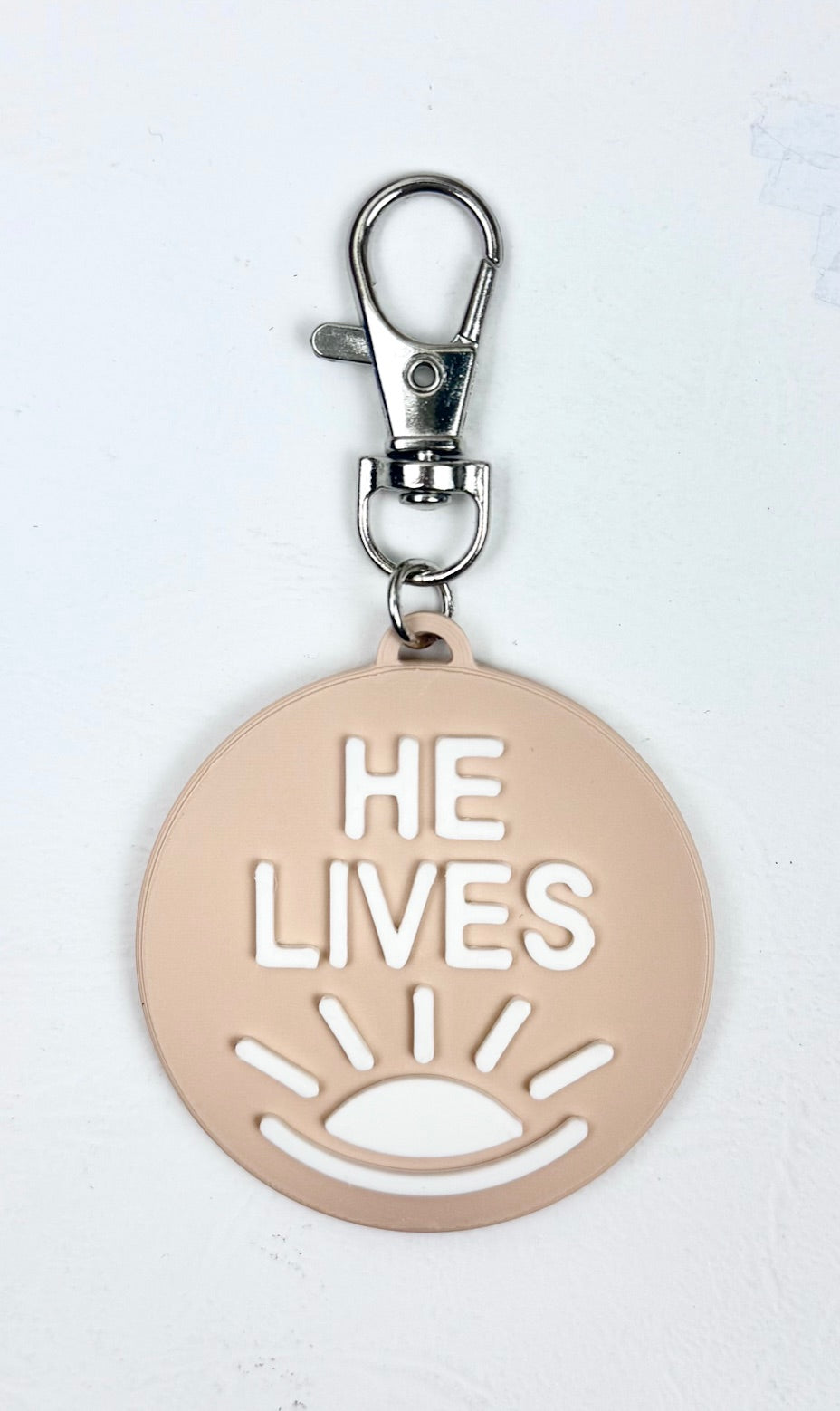Sand-colored bag charm with a lobster clasp and white 'He Lives' lettering and a rising sun icon