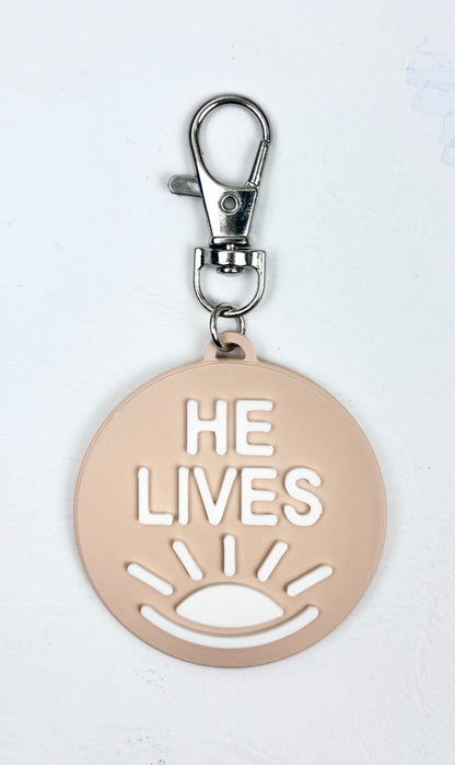 Sand-colored bag charm with a lobster clasp and white 'He Lives' lettering and a rising sun icon