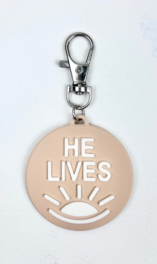 Sand-colored bag charm with a lobster clasp and white 'He Lives' lettering and a rising sun icon