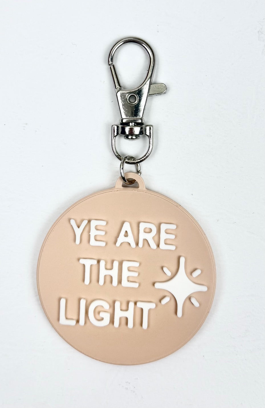 Sand-colored bag charm with a lobster clasp and white 'Ye Are the Light' lettering and shining star icon