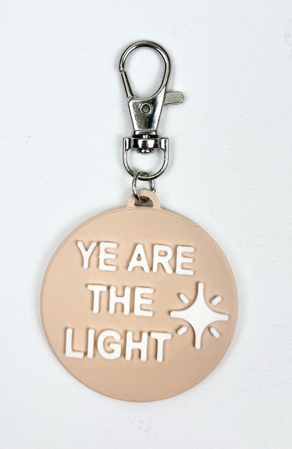 Sand-colored bag charm with a lobster clasp and white 'Ye Are the Light' lettering and shining star icon