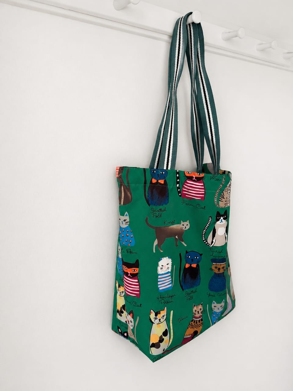 Green tote bag with cat pattern hanging on a white wall