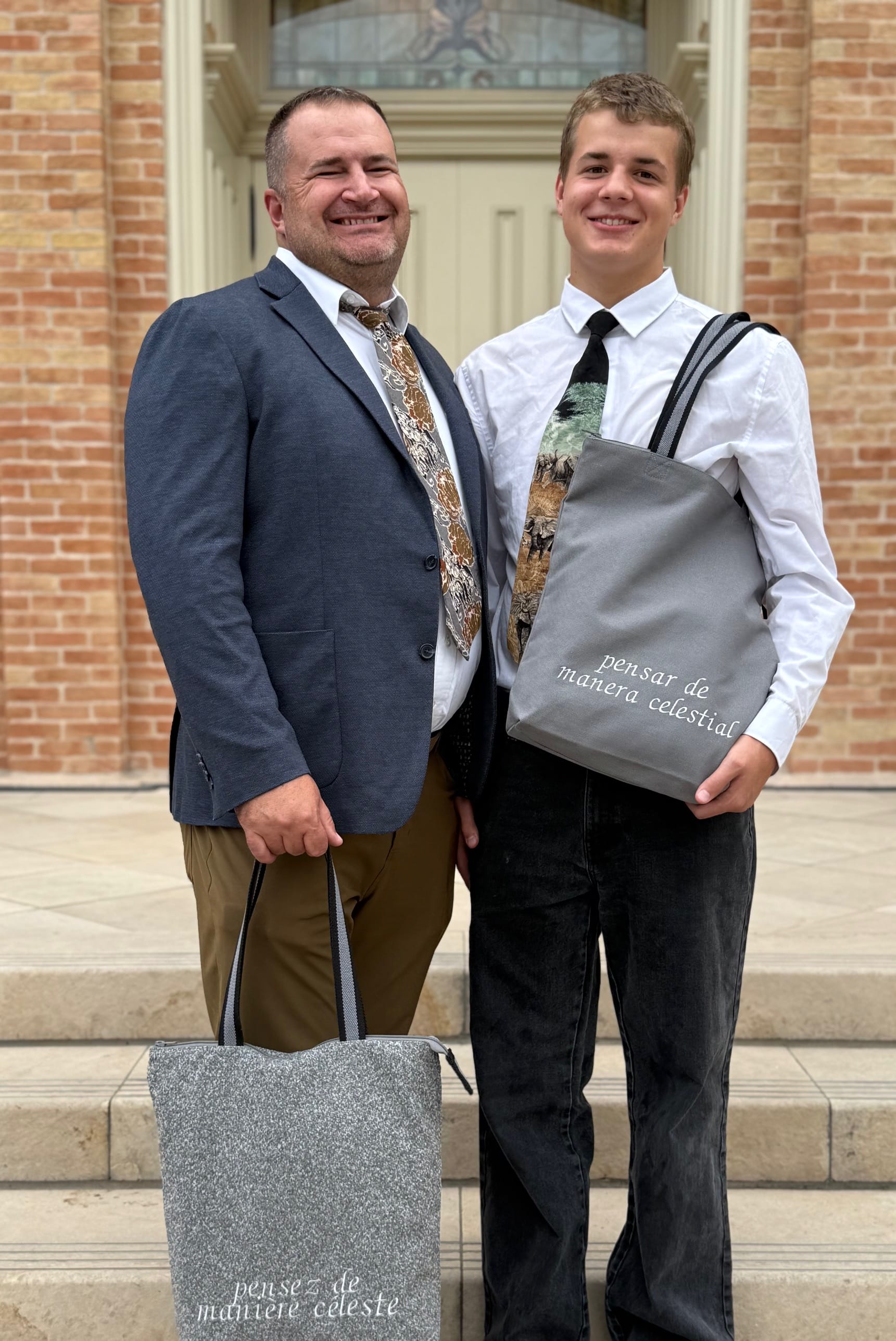 Father and son smiling on temple steps, holding temple bags with “pensez de manière céleste” and “pensar de manera celestial” embroidered on front