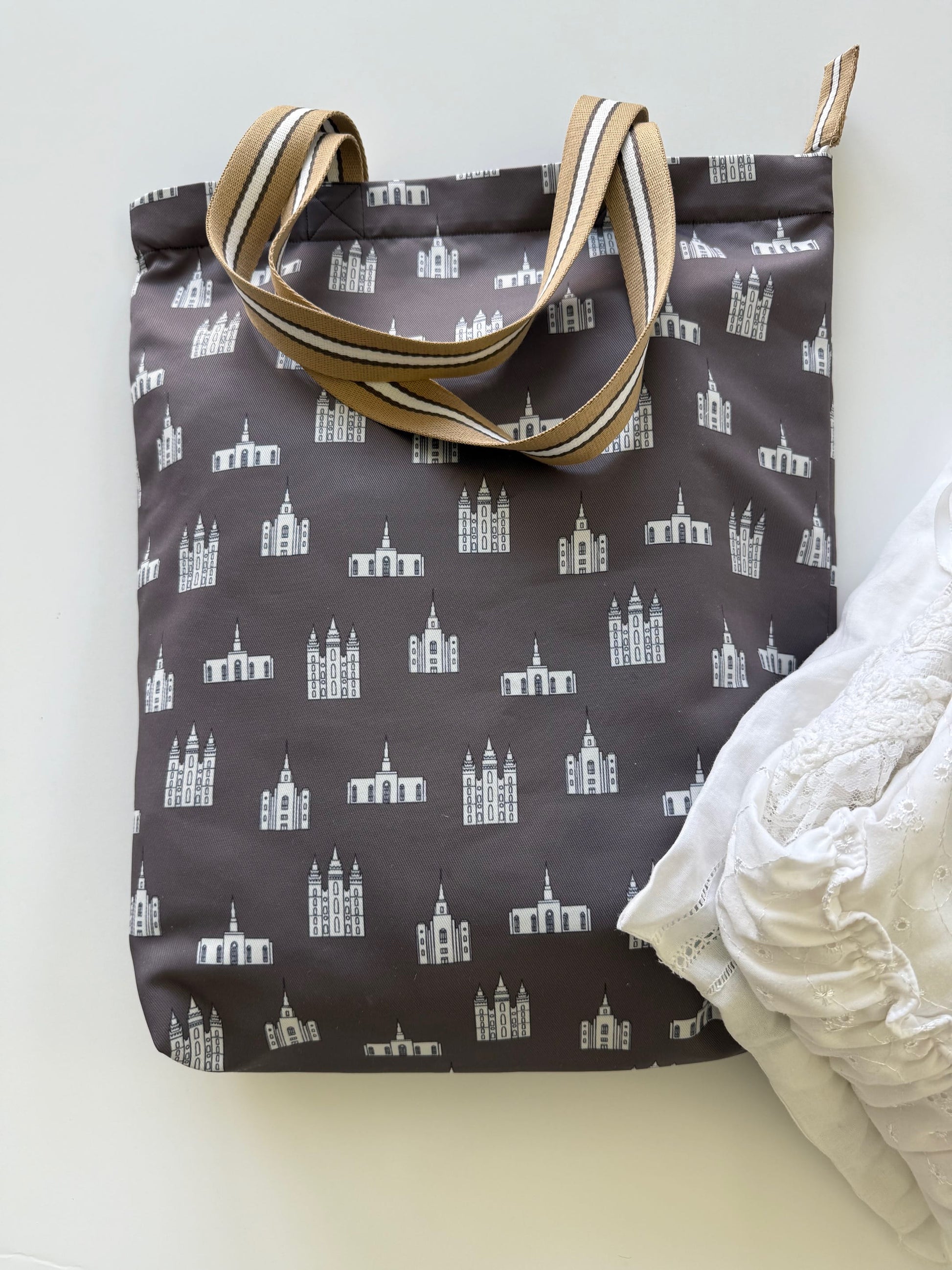 Brown LDS tote bag with white temple illustrations and tan striped handles, next to white folded temple clothing.