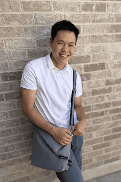 Man in a white polo shirt and gray pants leaning against a brick wall and carrying a gray crossbody shoulder bag