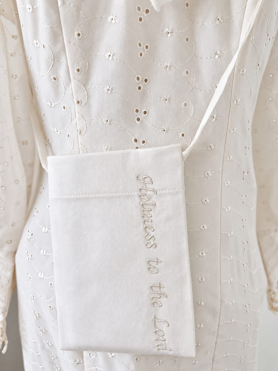 White cotton temple pouch embroidered with the phrase “Holiness to the Lord,” worn in front of a white eyelet temple dress.