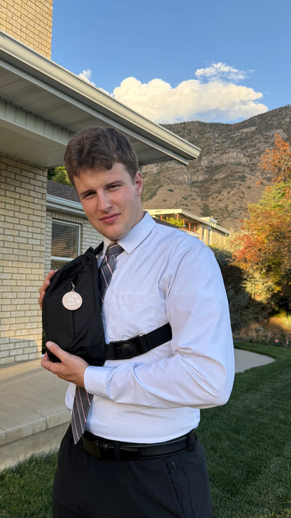 Angled view of the black LDS Mission Ready Convertible Bag worn crossbody, showing the strap buckle, front pocket, and bag charm