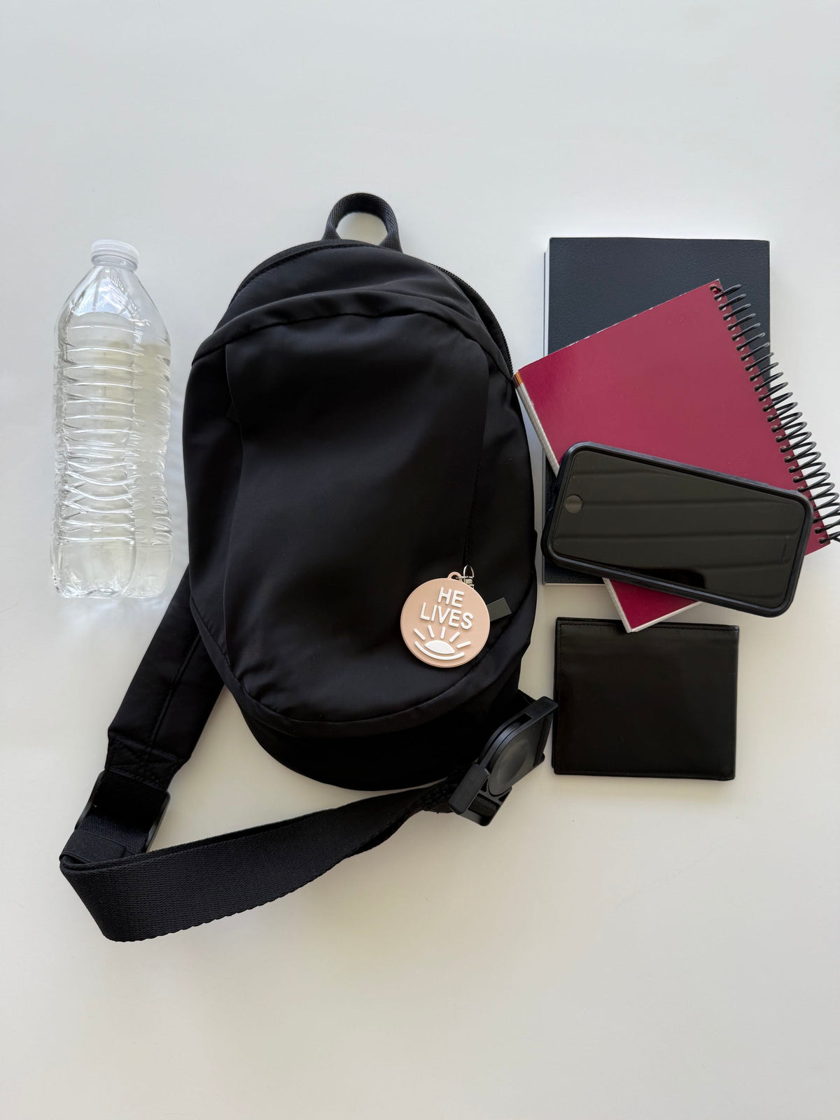 Black convertible bag with a “He Lives” charm beside a water bottle, book, spiral notebook, phone, and wallet