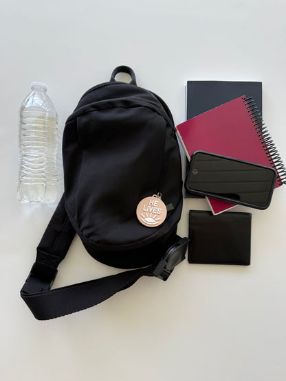 Black convertible bag with a “He Lives” charm beside a water bottle, book, spiral notebook, phone, and wallet
