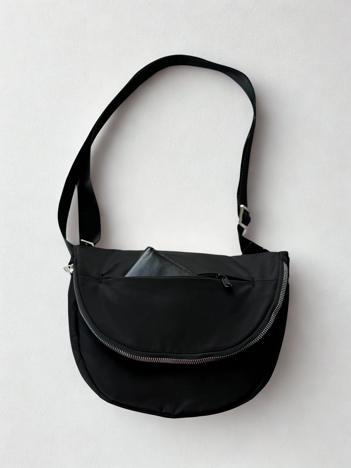 A black crossbody bag with a curved zipper closure and a long, adjustable strap. The bag has a minimalist design and is made of a durable, water-resistant material. A small, black wallet is tucked inside the outer pocket of the bag.