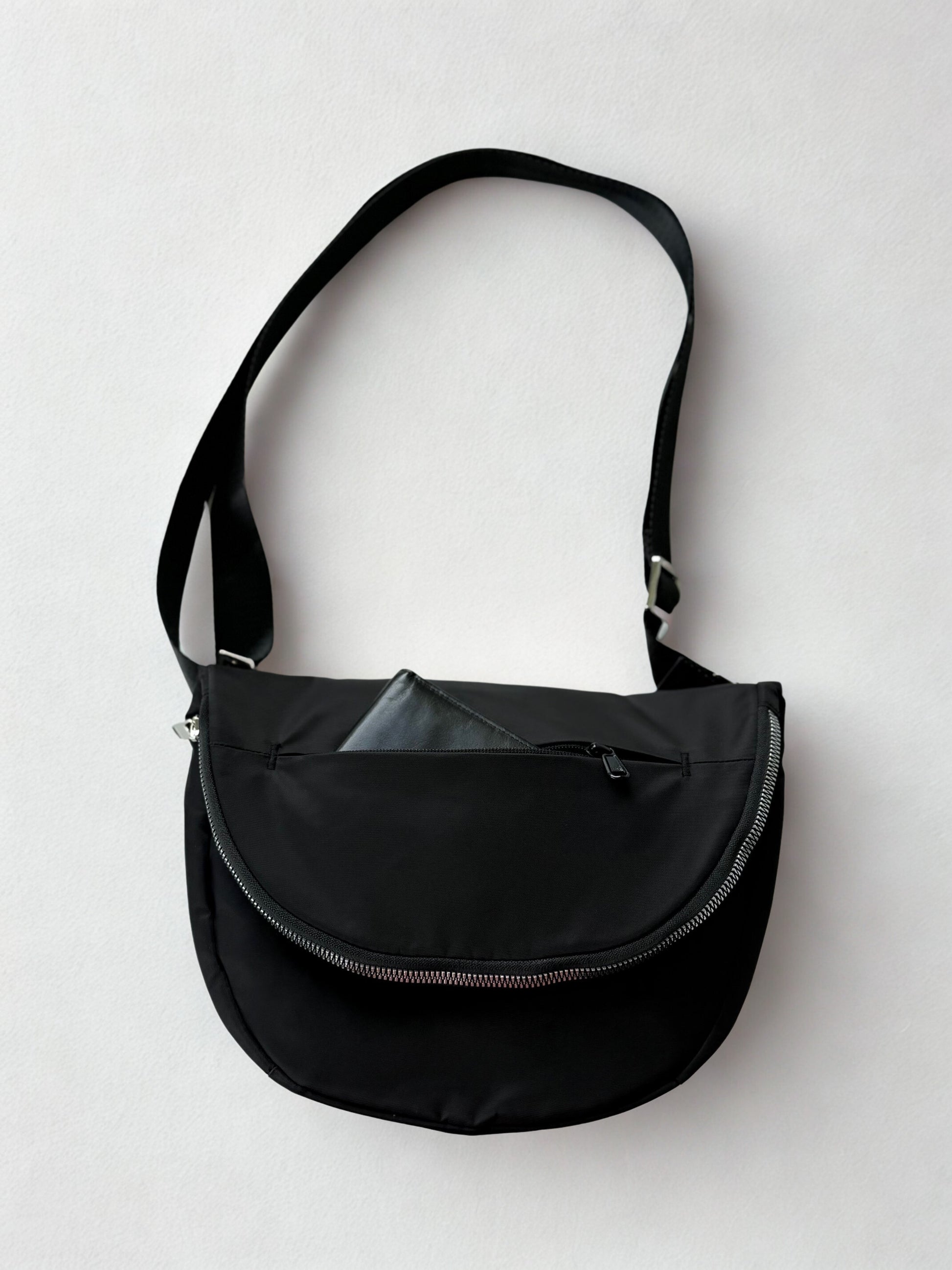 A black crossbody bag with a curved zipper closure and a long, adjustable strap. The bag has a minimalist design and is made of a durable, water-resistant material. A small, black wallet is tucked inside the outer pocket of the bag.