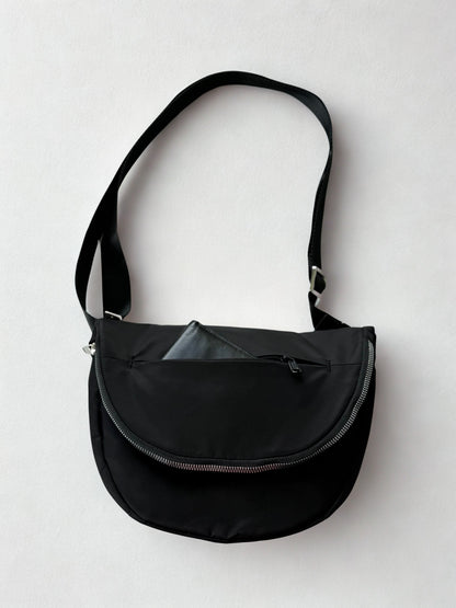 A black crossbody bag with a curved zipper closure and a long, adjustable strap. The bag has a minimalist design and is made of a durable, water-resistant material. A small, black wallet is tucked inside the outer pocket of the bag.