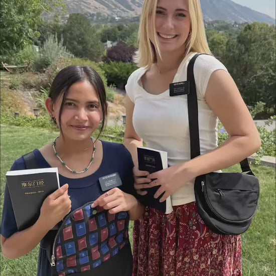 Two sister missionaries display black missionary bags outdoors, one open to reveal its colorful lining