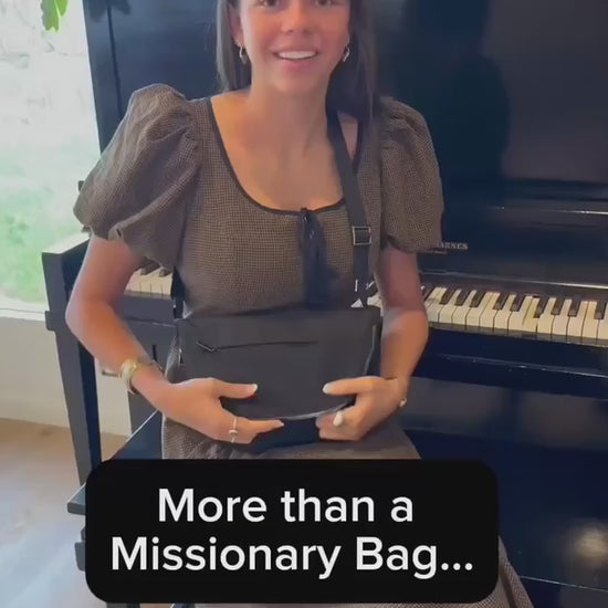 A young woman opens a black crossbody missionary bag to show its interior compartments and colorful scripture-patterned lining