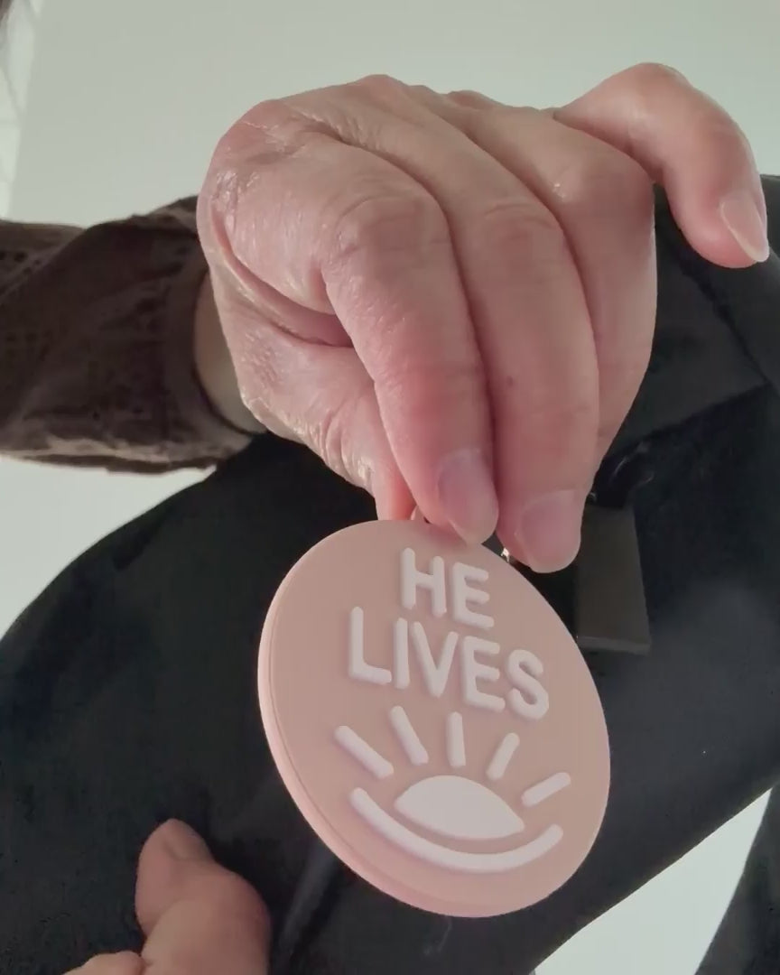 Demonstration of the two sides of the bag charm. One side with the words "He Lives and the other "Ye Are the Light"